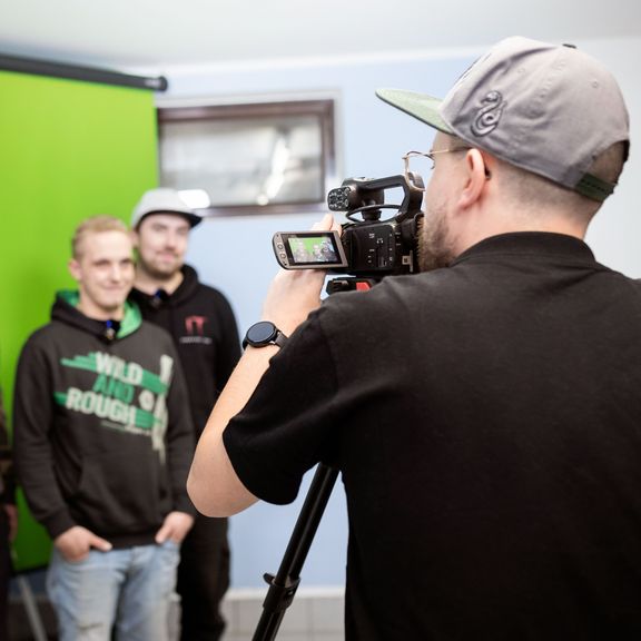 Drei junge Männer stehen vor einem Greenscreen und werden von einem Mann mit Videokamera gefilmt.