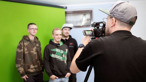 Drei junge Männer stehen vor einem Greenscreen und werden von einem Mann mit Videokamera gefilmt.