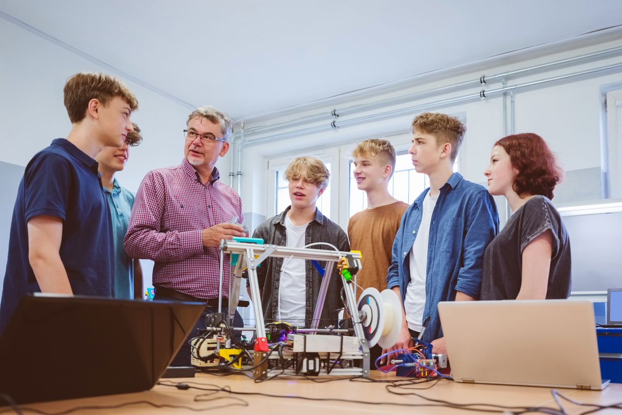Gruppe mit sechs Jugendlichen stehen in einem Raum zusammen, in der Mitte steht der Lehrer, der erklärt.