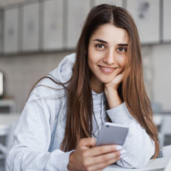 How to Azubirecruiting Eine junge Frau sitzt vor ihrem Laptop mit dem Smartphone in der Hand und lächelt in die Kamera.