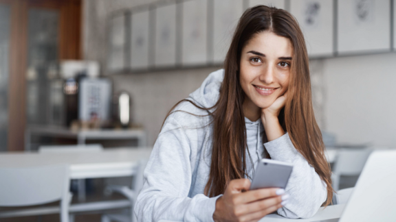 Eine junge Frau sitzt vor ihrem Laptop mit dem Smartphone in der Hand und lächelt in die Kamera.