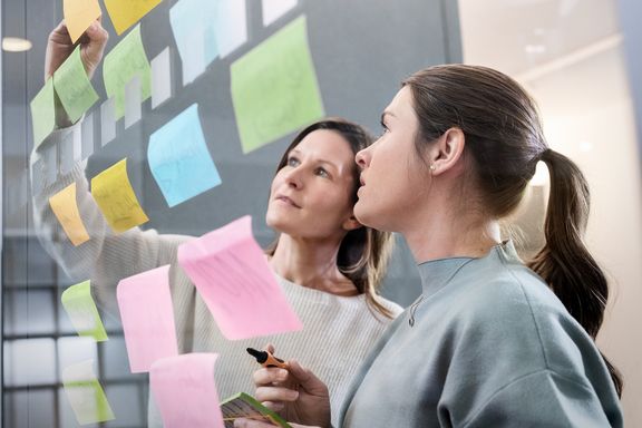 Zwei Frauen stehen vor einer Glaswand und kleben Post-It´s darauf