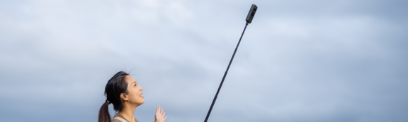 Frau mit Selfie-Stick vor bewölktem Himmel draußen.