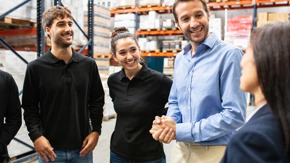 Eine Gruppe Mitarbeitender unterhält sich gut gelaunt in einem Lager mit hohen Regalen im Hintergrund.
