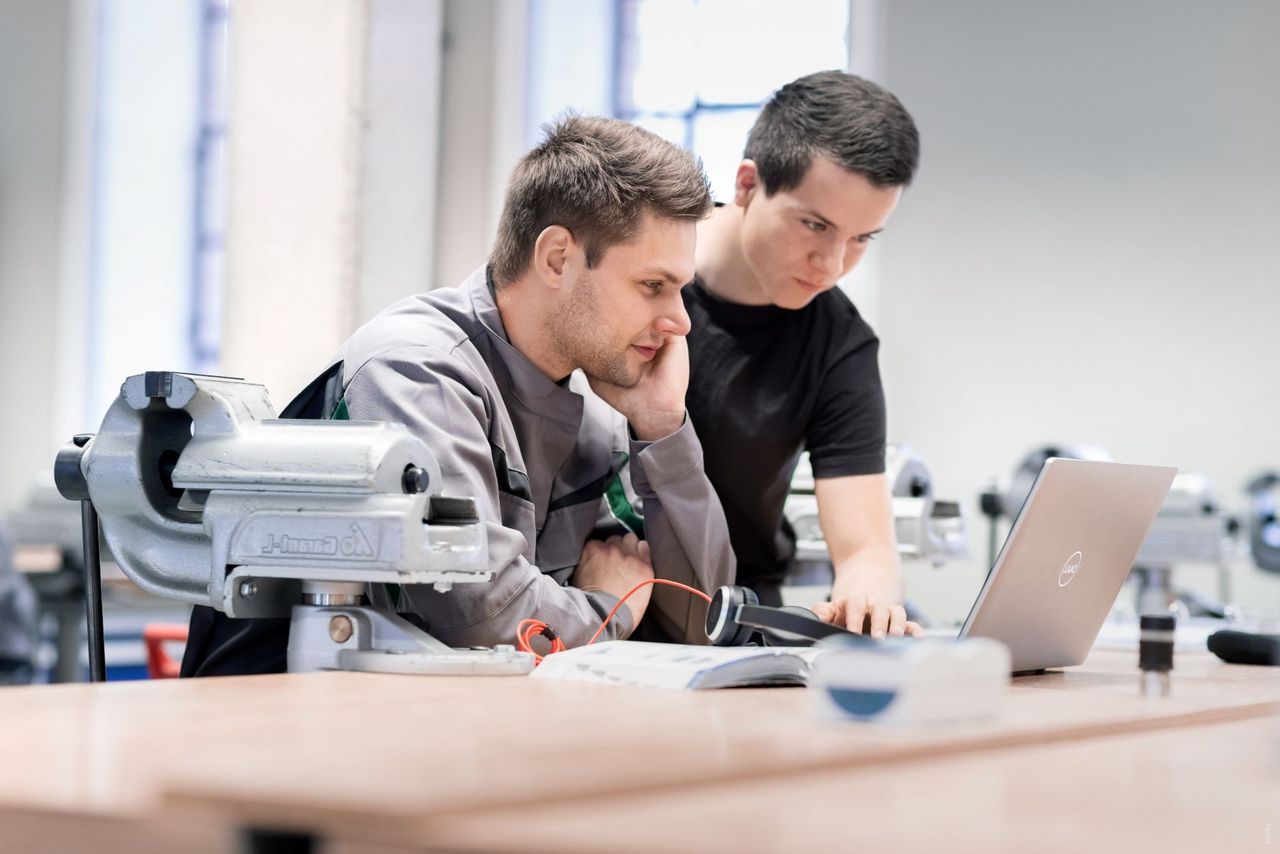 Ausbilder und Azubi sind in einer Werkstatt. Beide schauen freundlich auf den Bildschirm des Laptops