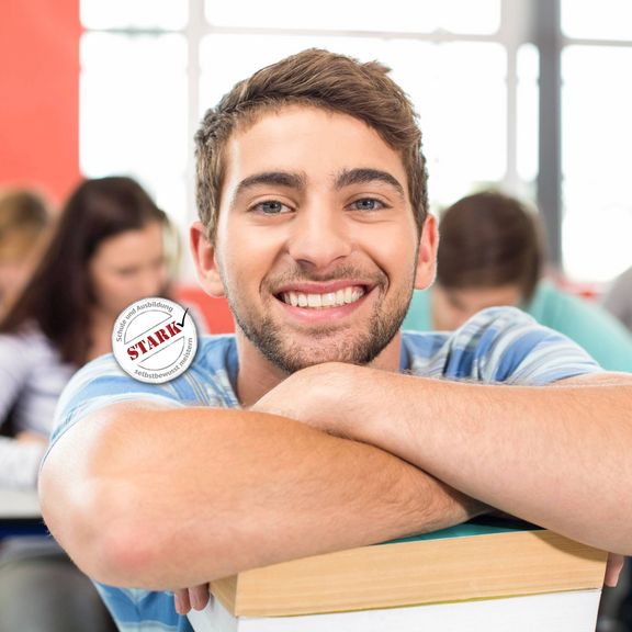 STARK Workshop für Resilienz und Stresskompetenz Ein junger Mann in einem Klassenzimmer verschränkt die Arme auf einem Stapel Bücher und lächelt in die Kamera.