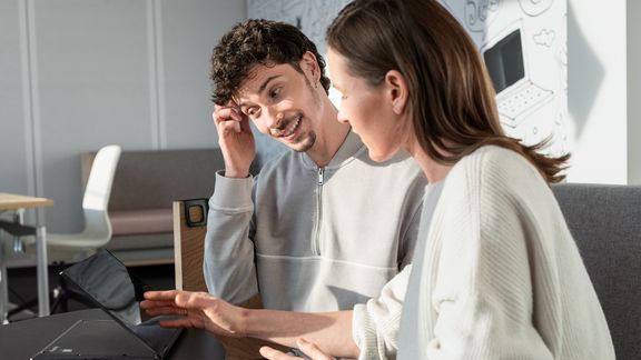Eine Frau und ein Mann, die nebeneinander sitzen. Die Frau erklärt dem Mann etwas, und der Mann überlegt und grübelt.