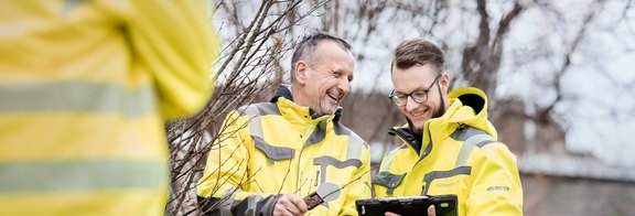 Zwei Männer, in gelber Arbeitskleidung, schauen gemeinsam auf ein Tablet.