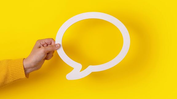 Eine Hand hält vom linken Rand eine weiße Sprechblase in das Bild vor einem gelben Hintergrund.