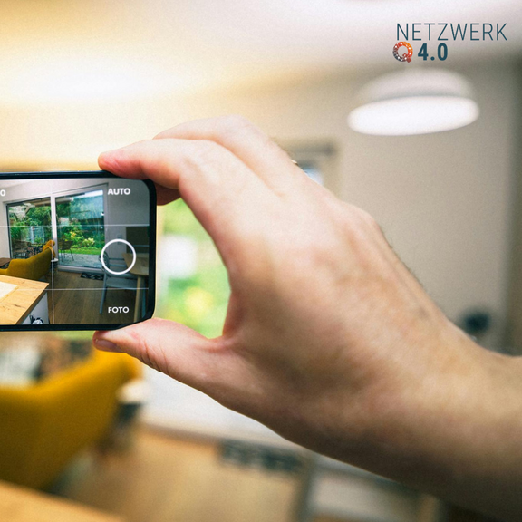 Hand hält Smartphone mit geöffneter Kamera-App in einem hellen Wohnzimmer, Logo „Netzwerk 4.0“.