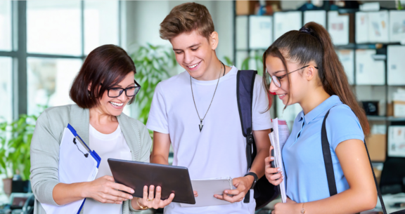 Eine Frau zeigt zwei lächelnden Jugendlichen in einem hellen Büro oder Klassenzimmer etwas auf einem Tablet, während alle gemeinsam darauf schauen.