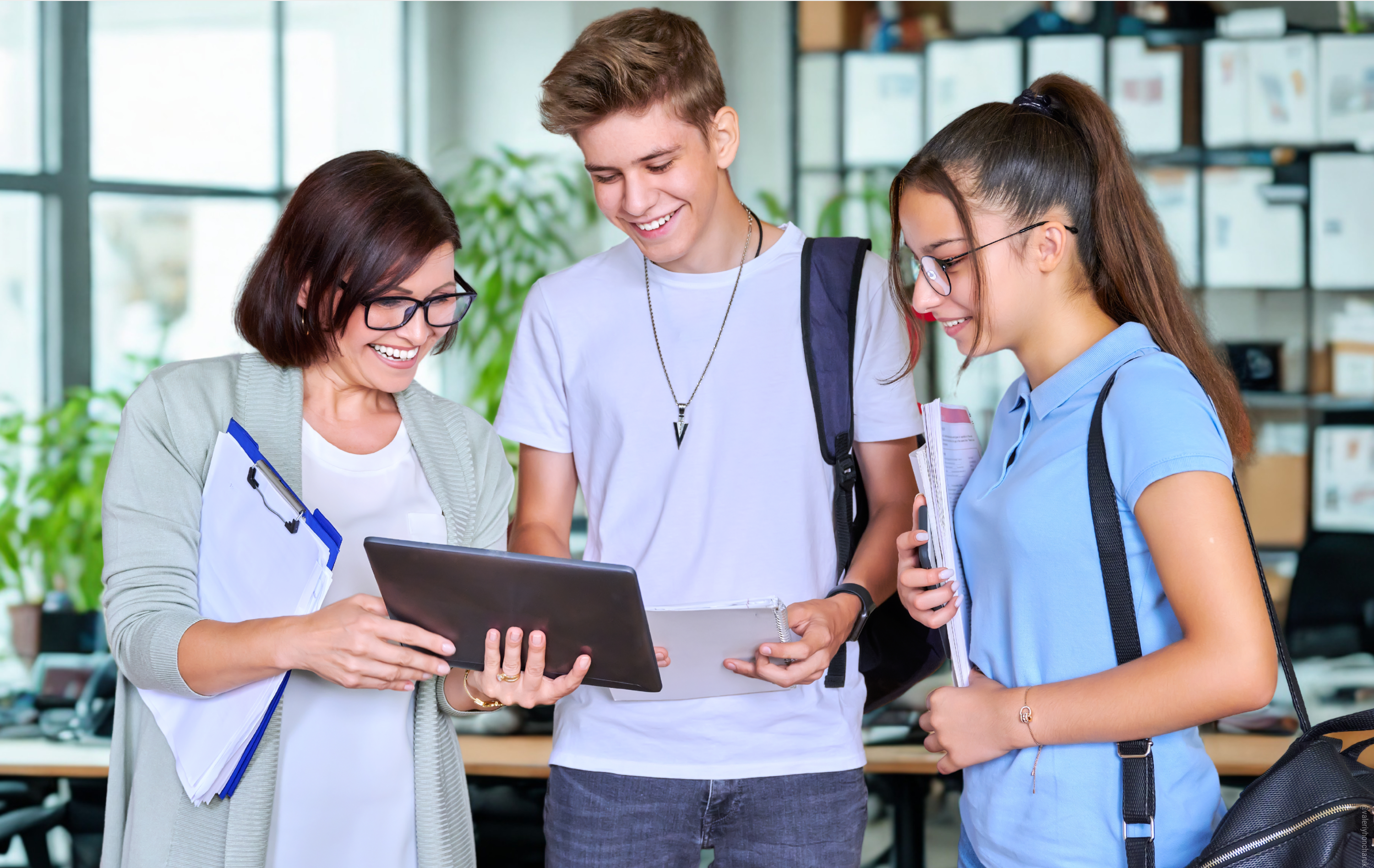 Eine Frau zeigt zwei lächelnden Jugendlichen in einem hellen Büro oder Klassenzimmer etwas auf einem Tablet, während alle gemeinsam darauf schauen.